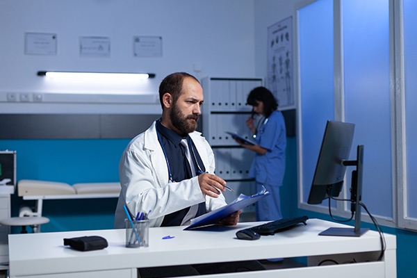 A doctor sitting at a computer looking at it with a clipboard in his hand with a nurse in the room in the background