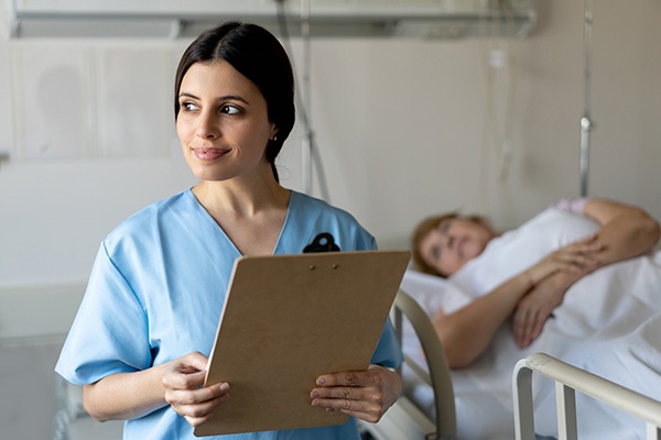 A nurse with a clipboard in her hand while a patient lays on a hospital bed in the background