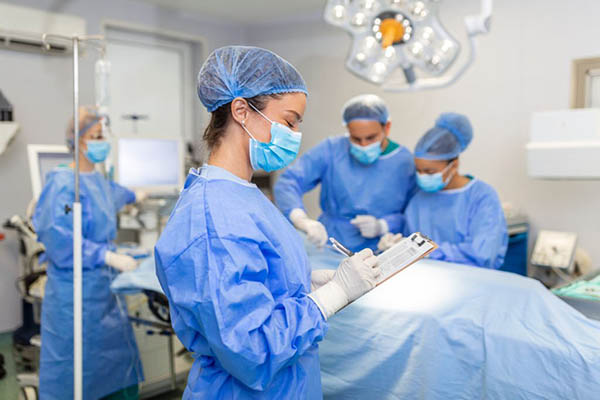 A CRNA making notes on a clipboard with a surgery going on in the background