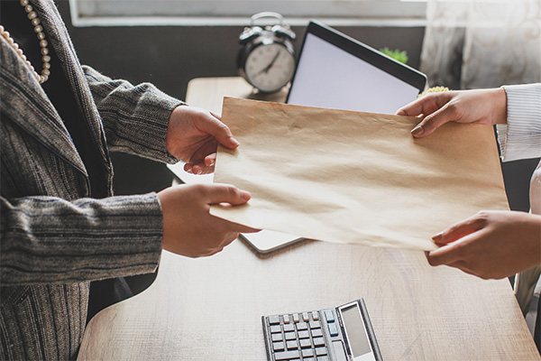 A person handing a sheet of paper to another person standing over top of a desk with a laptop in the background