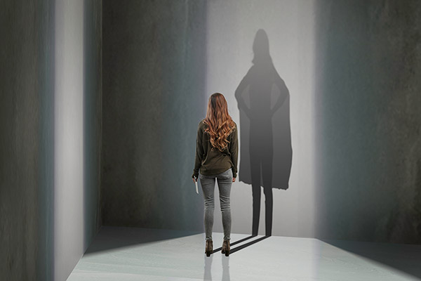 A woman standing in a room with her shadow cast upon the wall-. The shadow is made to look like a superhero. 