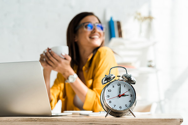 A woman holding a cup of coffee sitting at a laptop with an alarm clock nearby