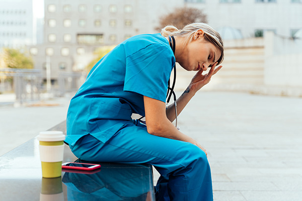 A nurse sitting on a bench outside with her head in her hands looking stressed. 