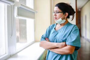 A nurse looking out a window while standing in a hospital hallways. They have their arms crossed in front of them. 